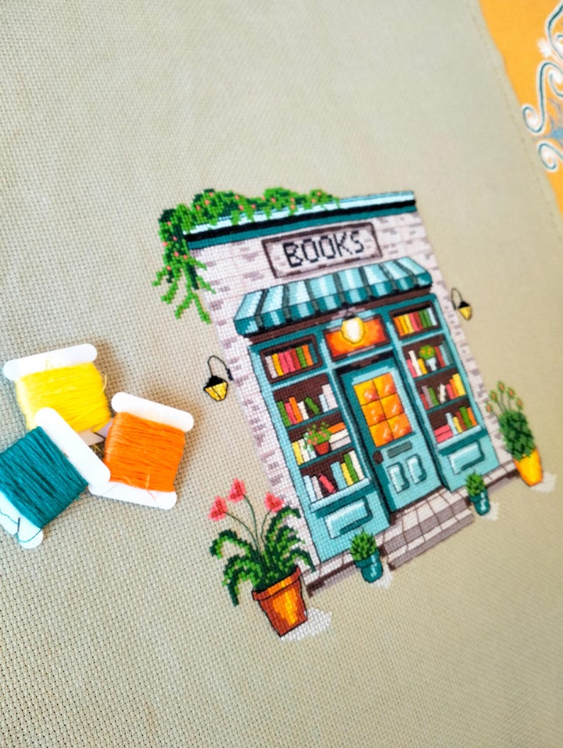 Op de afbeelding: Een kruissteekborduurwerk op lichtbeige stof met een boekwinkel met een blauwgroene pui, een gestreepte luifel en het woord BOOKS boven de deur. Drie klossen borduurgaren in geel, blauwgroen en oranje liggen aan de linkerkant.