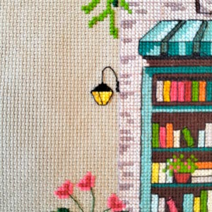 Op de afbeelding: Een kruissteekborduurwerk op beige stof met een winkelpui met een blauwgroen frame, een gestreepte luifel en planken vol kleurrijke boeken. Een zwarte wandlamp, groene hangende klimplanten en roze bloemen zijn links geborduurd.