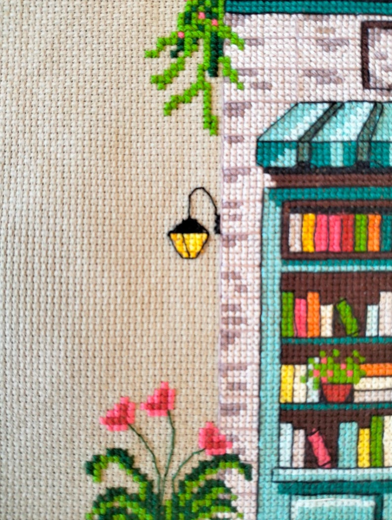 Op de afbeelding: Een kruissteekborduurwerk op beige stof met een winkelpui met een blauwgroen frame, een gestreepte luifel en planken vol kleurrijke boeken. Een zwarte wandlamp, groene hangende klimplanten en roze bloemen zijn links geborduurd.