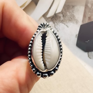 Silver ring with a cowrie shell held by a hand on a textured surface.