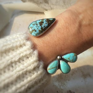 May include: Two silver bracelets with turquoise stones. One bracelet features a teardrop-shaped stone with blue and brown patterns. The other bracelet has three turquoise stones in a floral design. The bracelets are worn on a wrist.