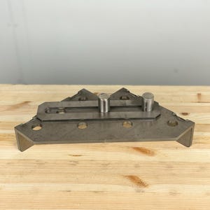 K&ouml;nnte beinhalten: Eine Metall-Eckklemme mit zwei verstellbaren Stiften. Die Klemme ist aus Stahl gefertigt und hat eine graue Oberfl&auml;che. Sie ist so konzipiert, dass sie zwei Holzst&uuml;cke in einem 90-Grad-Winkel zusammenh&auml;lt.