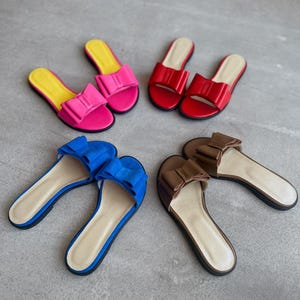 May include: Four pairs of flat slide sandals arranged in a circle. The sandals feature a prominent bow on the strap and come in four distinct colors: bright pink with a yellow insole, solid red, vibrant blue, and a muted brown with a beige insole.