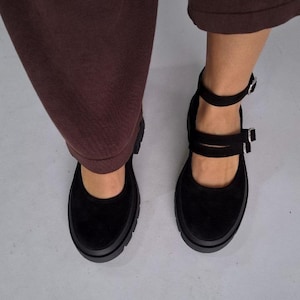 Op de afbeelding: Zwarte Mary Jane-schoenen met een dikke zool en dubbele enkelbandjes. De schoenen hebben een suède-achtige textuur en zilveren gespen. De schoenen worden gedragen met een bruine broek.