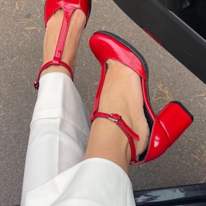 May include: A pair of red patent leather heeled shoes with a T-strap and ankle strap. The shoes have a chunky heel.