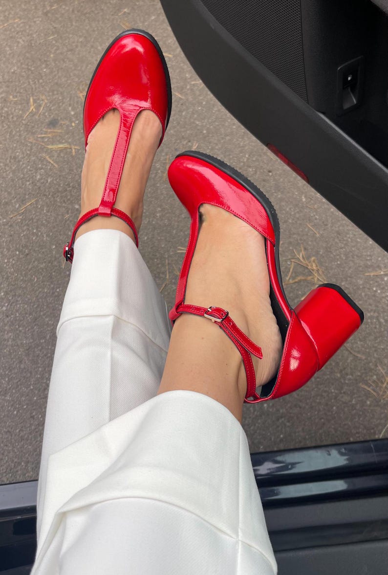 May include: A pair of red patent leather heeled shoes with a T-strap and ankle strap. The shoes have a chunky heel.
