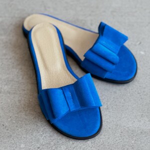 May include: A pair of bright blue open-toe slide sandals featuring a large, structured bow detail on the top strap. The footbed is a light beige color, and the sole is thin and black, providing a contrast to the vibrant blue upper material.