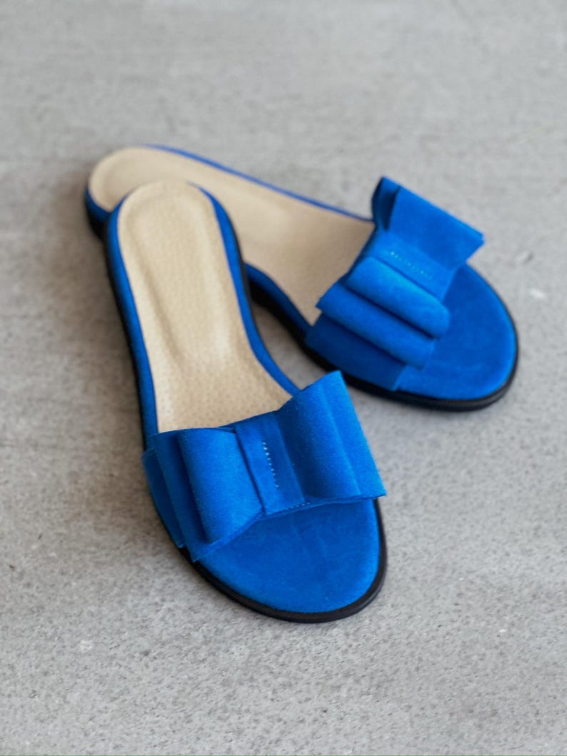 May include: A pair of bright blue open-toe slide sandals featuring a large, structured bow detail on the top strap. The footbed is a light beige color, and the sole is thin and black, providing a contrast to the vibrant blue upper material.