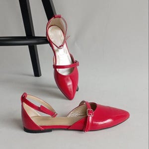 Peut inclure: Une paire de chaussures plates rouge vif à bout pointu avec une finition vernie brillante. Chaque chaussure présente une fine bride de cheville avec une petite boucle en métal et une seconde bride sur le cou-de-pied. L'intérieur est beige.