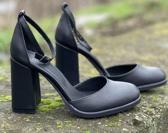 Op de afbeelding: Een paar zwarte leren hakken met enkelbandjes. De schoenen hebben een dikke blokhak en een afgeronde neus.