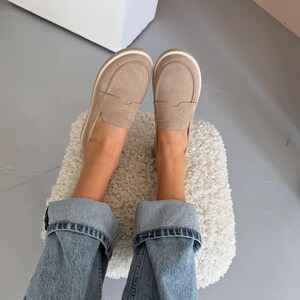 May include: A pair of light brown suede loafers with a white sole and trim. The shoes are worn with blue denim jeans. The image is taken from a top-down perspective.