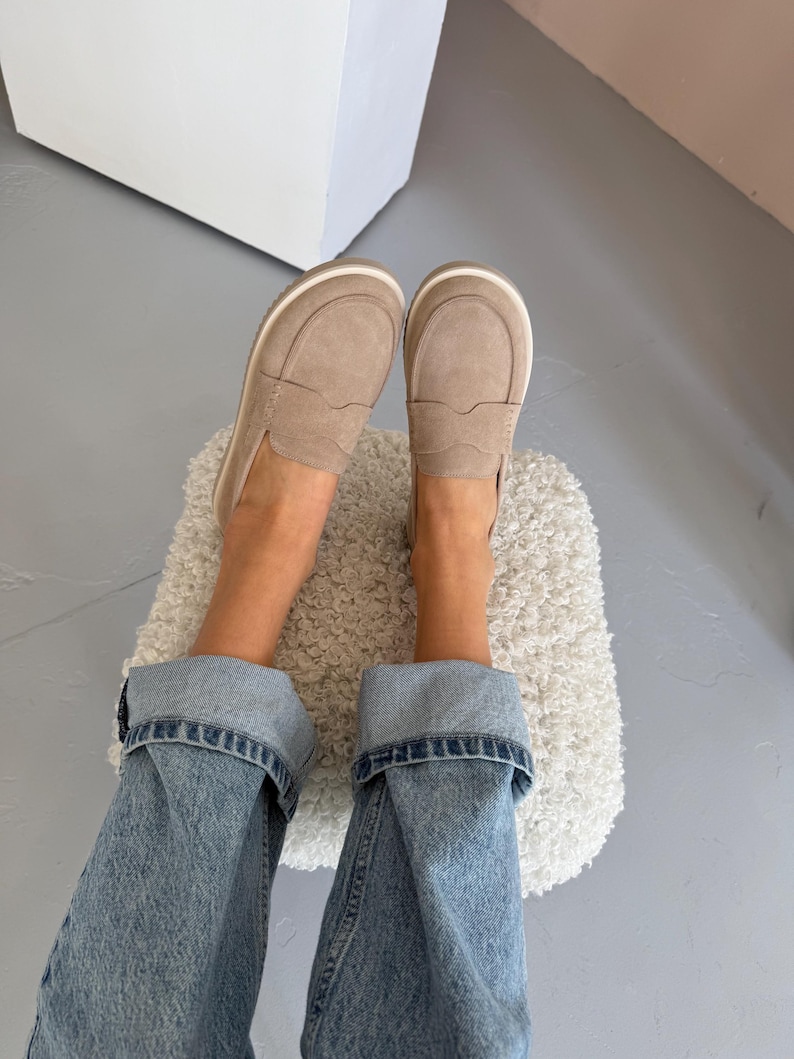 May include: A pair of light brown suede loafers with a white sole and trim. The shoes are worn with blue denim jeans. The image is taken from a top-down perspective.
