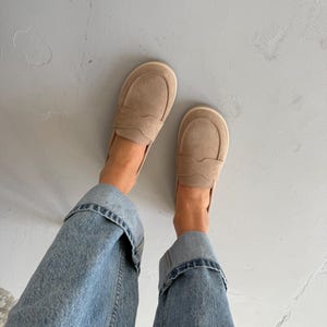 May include: A pair of beige suede loafers with a rounded toe and decorative stitching. The shoes are paired with light blue denim jeans with rolled-up cuffs. The loafers are a neutral color, suitable for various outfits.