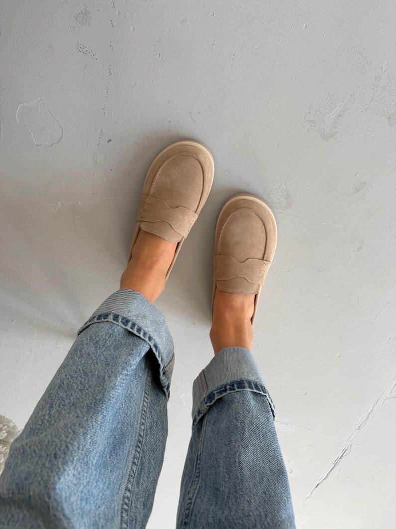 May include: A pair of beige suede loafers with a rounded toe and decorative stitching. The shoes are paired with light blue denim jeans with rolled-up cuffs. The loafers are a neutral color, suitable for various outfits.