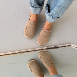 May include: A pair of tan suede loafers with a rounded toe and a decorative stitched design. The shoes are worn with light blue denim jeans with rolled-up cuffs. The image is taken from a high-angle perspective.