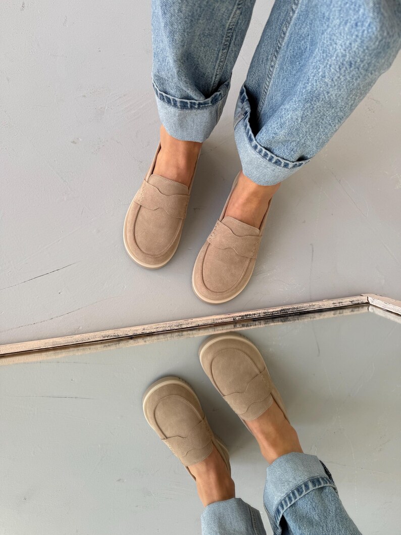 May include: A pair of tan suede loafers with a rounded toe and a decorative stitched design. The shoes are worn with light blue denim jeans with rolled-up cuffs. The image is taken from a high-angle perspective.