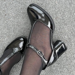 May include: A pair of black patent leather heeled shoes with a strap and buckle detail. The shoes have a mesh panel on the front and are worn with black tights.