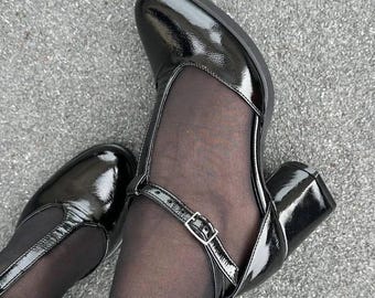 May include: A pair of black patent leather heeled shoes with a strap and buckle detail. The shoes have a mesh panel on the front and are worn with black tights.