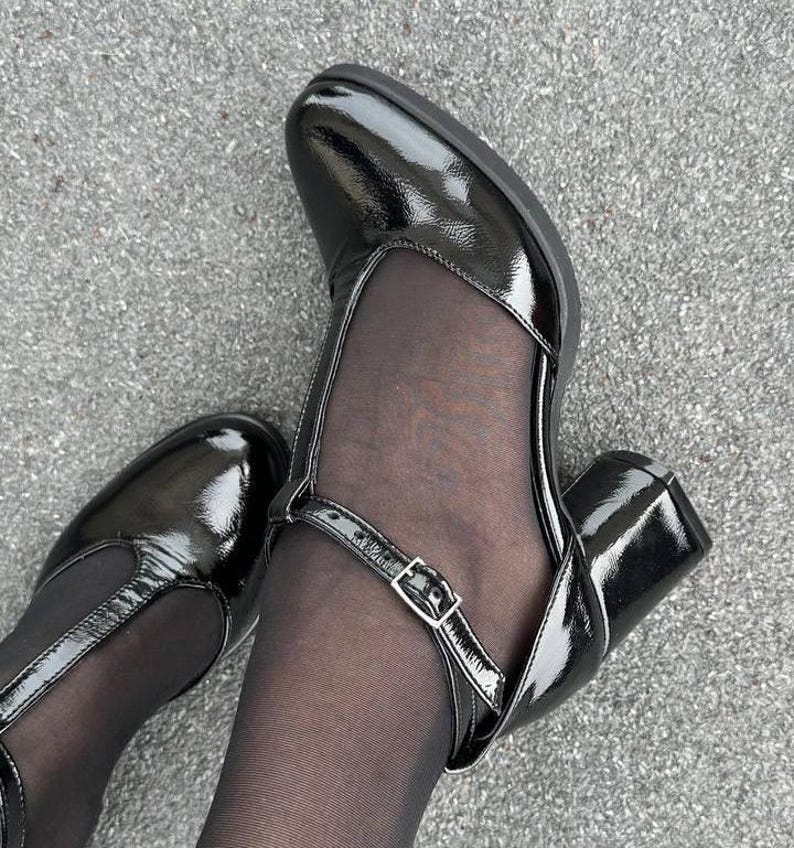 May include: A pair of black patent leather heeled shoes with a strap and buckle detail. The shoes have a mesh panel on the front and are worn with black tights.