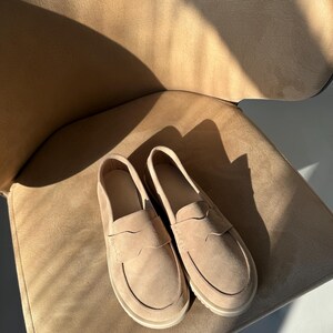 May include: A pair of beige suede loafers with a classic penny loafer design. The shoes have a rounded toe and a low, off-white sole. They are placed on a beige chair, with sunlight casting shadows.