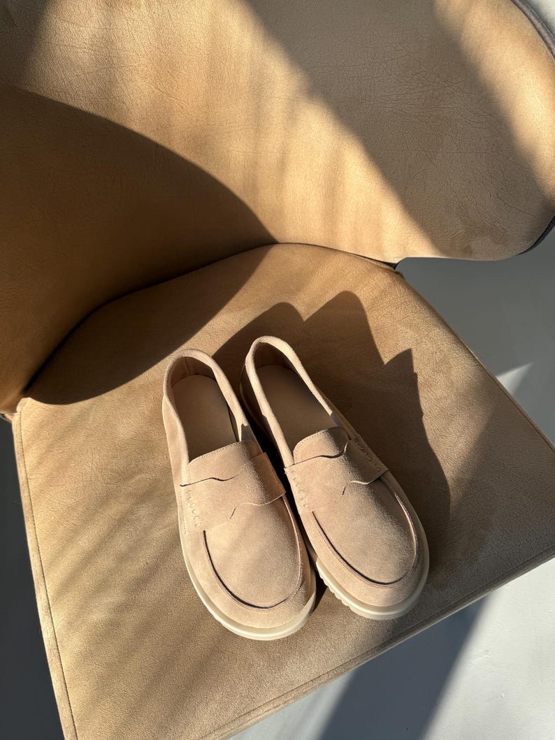 May include: A pair of beige suede loafers with a classic penny loafer design. The shoes have a rounded toe and a low, off-white sole. They are placed on a beige chair, with sunlight casting shadows.