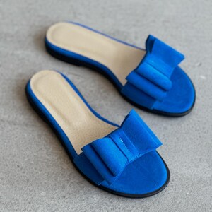 May include: A pair of bright blue flat sandals featuring a large, structured bow detail on the top strap. The sandals have a light beige insole and a thin black outsole, providing a simple slip-on design with a vibrant, solid color finish.