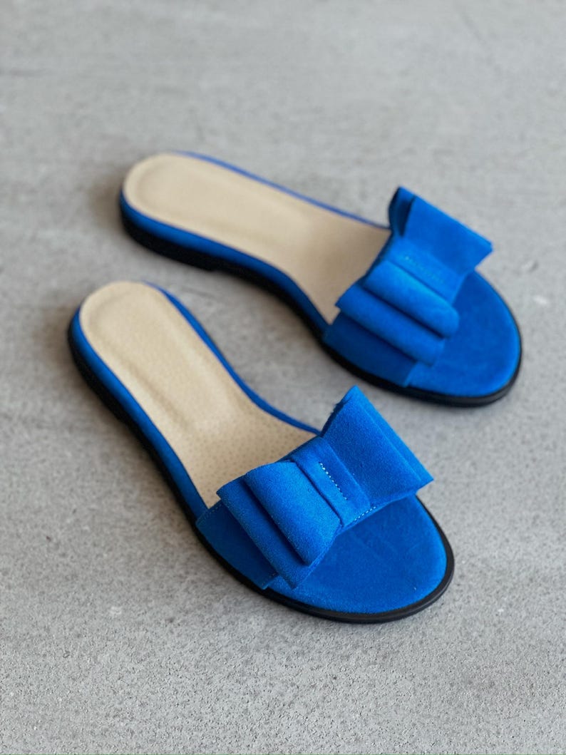 May include: A pair of bright blue flat sandals featuring a large, structured bow detail on the top strap. The sandals have a light beige insole and a thin black outsole, providing a simple slip-on design with a vibrant, solid color finish.