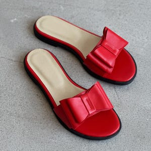 May include: A pair of flat slide sandals in vibrant red, featuring a prominent decorative bow on the wide front strap. The footbed is a light beige color, and the outsole is black. These open-toe slip-on sandals have a simple, low-profile design.