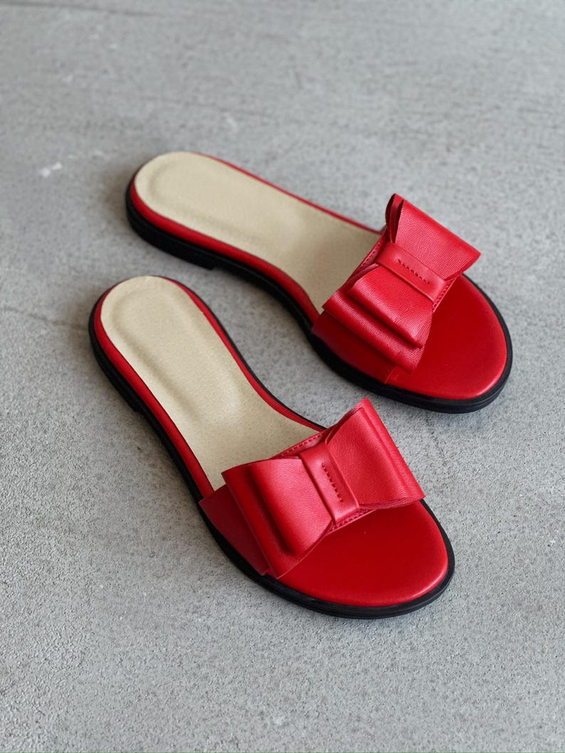 May include: A pair of flat slide sandals in vibrant red, featuring a prominent decorative bow on the wide front strap. The footbed is a light beige color, and the outsole is black. These open-toe slip-on sandals have a simple, low-profile design.