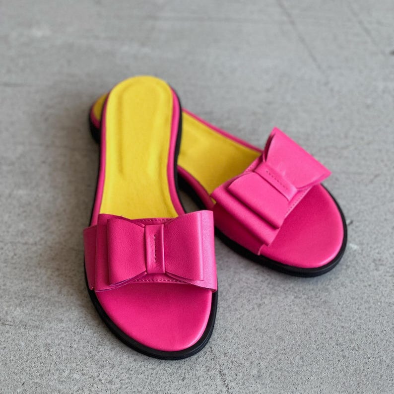 May include: A pair of slide sandals featuring a vibrant hot pink upper with a prominent decorative bow on the strap. The footbed is a bright yellow color, and the sole is outlined in black, providing a distinct contrast to the pink and yellow.