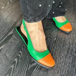 Könnte beinhalten: Ein Paar grüne und orangefarbene Lederschuhe mit Absatz und Slingback-Riemen und einer Schnalle. Die Schuhe stehen auf einem Holzboden.