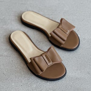 May include: A pair of brown slide sandals featuring a prominent bow detail on the upper strap. The sandals have a light beige footbed and a thin black sole, providing a simple and functional design for casual footwear with a flat profile.