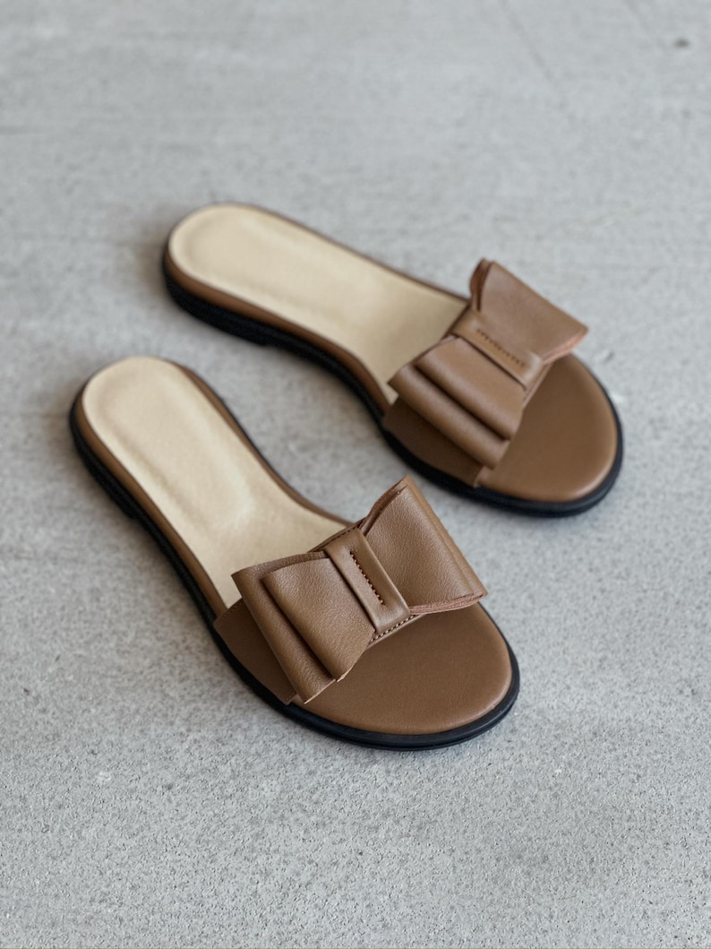 May include: A pair of brown slide sandals featuring a prominent bow detail on the upper strap. The sandals have a light beige footbed and a thin black sole, providing a simple and functional design for casual footwear with a flat profile.