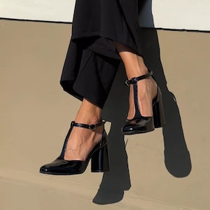 May include: Black patent leather T-strap heels with a block heel. The shoes have a pointed toe and an ankle strap with a buckle closure. The heels are approximately 3 inches high.