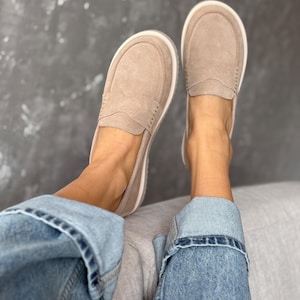 May include: A pair of tan suede loafers with white soles. The shoes have a classic penny loafer design with a stitched vamp and a rounded toe. The shoes are shown with blue denim jeans.