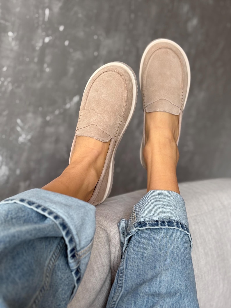May include: A pair of tan suede loafers with white soles. The shoes have a classic penny loafer design with a stitched vamp and a rounded toe. The shoes are shown with blue denim jeans.