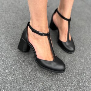 May include: Black leather heeled shoes with a T-strap and buckle closure. The shoes have a block heel and a pointed toe.