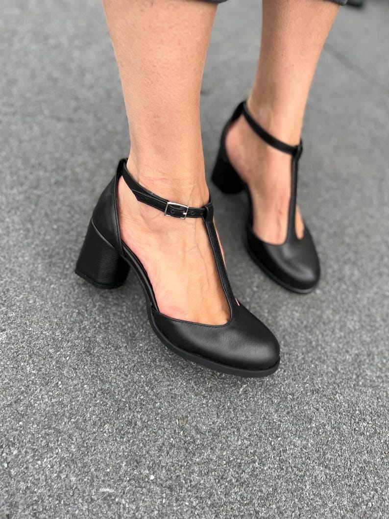 May include: Black leather heeled shoes with a T-strap and buckle closure. The shoes have a block heel and a pointed toe.