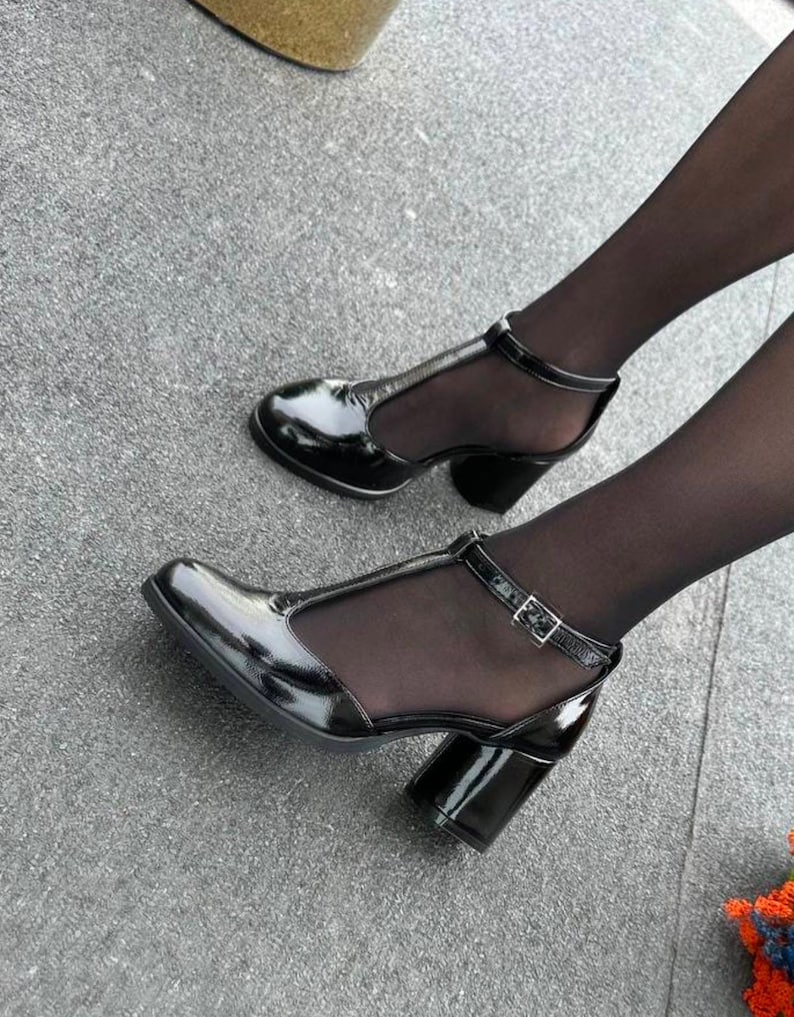 May include: Black patent leather heeled shoes with a T-strap and a mesh panel. The shoes have a chunky heel and a buckle closure.