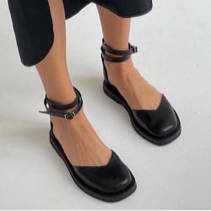 May include: Black leather sandals with a square toe and ankle straps. The sandals have a flat sole and a buckle closure. The straps wrap around the ankle and are secured with a small buckle. The shoes are a solid black color.