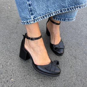 Könnte beinhalten: Schwarze Leder-Knöchelriemen-Heels mit Schleifendetail an der Spitze. Die Absätze haben ein Blockabsatz-Design. Die Schuhe werden mit blauen Jeans getragen.