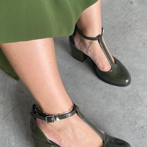 May include: A pair of green patent leather heeled shoes with a T-strap design. The shoes have a block heel and an ankle strap with a buckle closure.