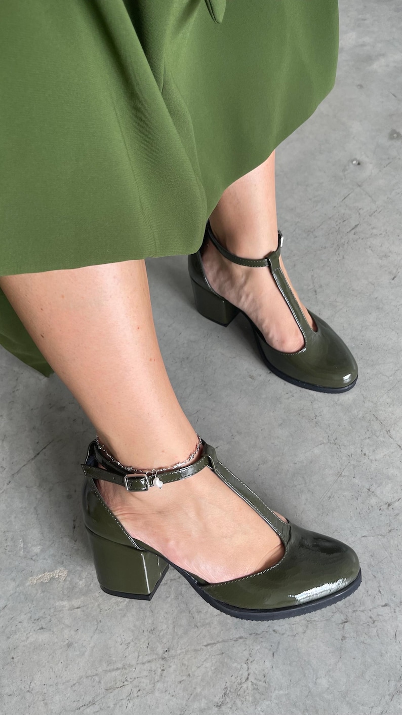 May include: A pair of green patent leather heeled shoes with a T-strap design. The shoes have a block heel and an ankle strap with a buckle closure.
