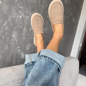May include: A pair of light brown suede slip-on shoes with white soles. The shoes are paired with blue denim jeans. The person is relaxing on a gray sofa against a gray textured wall.