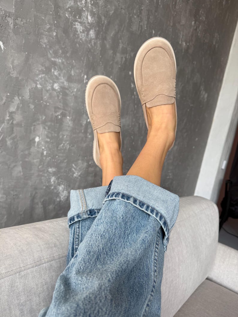 May include: A pair of light brown suede slip-on shoes with white soles. The shoes are paired with blue denim jeans. The person is relaxing on a gray sofa against a gray textured wall.