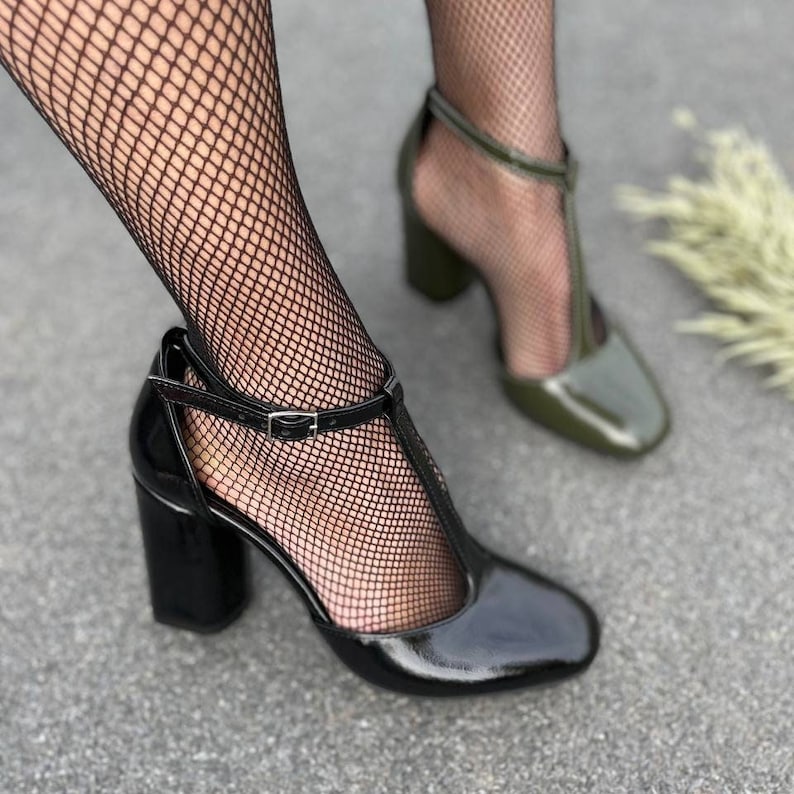 May include: A pair of high-heeled shoes with T-strap detailing. One shoe is black patent leather and the other is green patent leather.