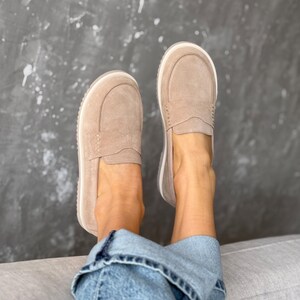 May include: A pair of light brown suede loafers with a white sole. The shoes have a classic slip-on design with a stitched vamp and a rounded toe. The shoes are shown on a person wearing blue jeans.