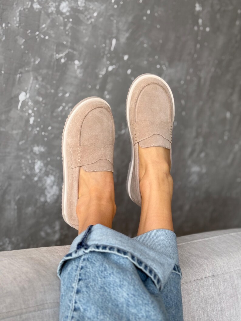 May include: A pair of light brown suede loafers with a white sole. The shoes have a classic slip-on design with a stitched vamp and a rounded toe. The shoes are shown on a person wearing blue jeans.