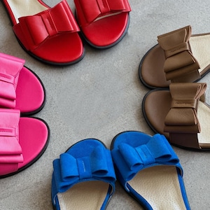 May include: A collection of three pairs of flat slide sandals arranged in a circle. Each pair features a prominent decorative bow on the strap. The sandals are available in vibrant red, bright pink, deep blue, and a neutral brown color palette.