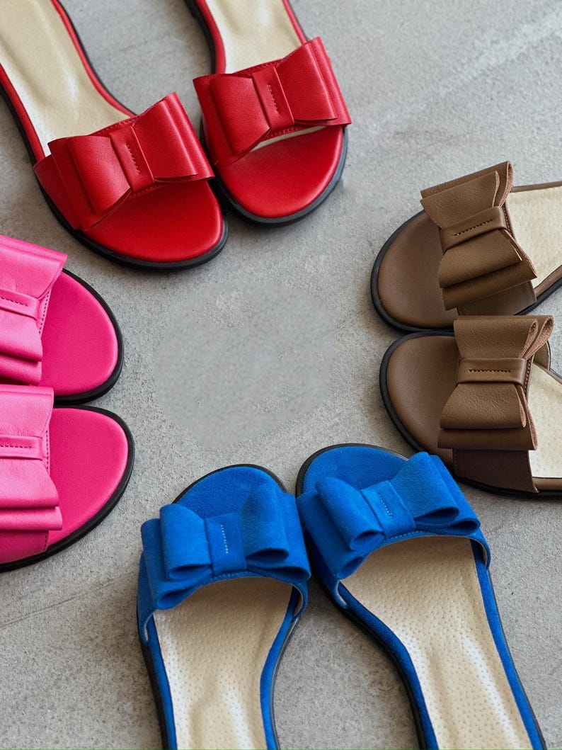 May include: A collection of three pairs of flat slide sandals arranged in a circle. Each pair features a prominent decorative bow on the strap. The sandals are available in vibrant red, bright pink, deep blue, and a neutral brown color palette.